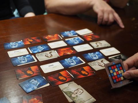 A game of Codenames at the end of play, played at restaurant Kaisla in Kaisaniemi, Helsinki, Finland. The game has ended because the assassin (the black card at the lower right corner of the grid) has been found. The key card, kept secret from the field operatives, can be seen at the extreme lower right corner of the picture, held up by one of the spymasters. The grid shown on the keycard corresponds to the actual grid of cards. The blue lights on the edges denote that the blue team started the game.